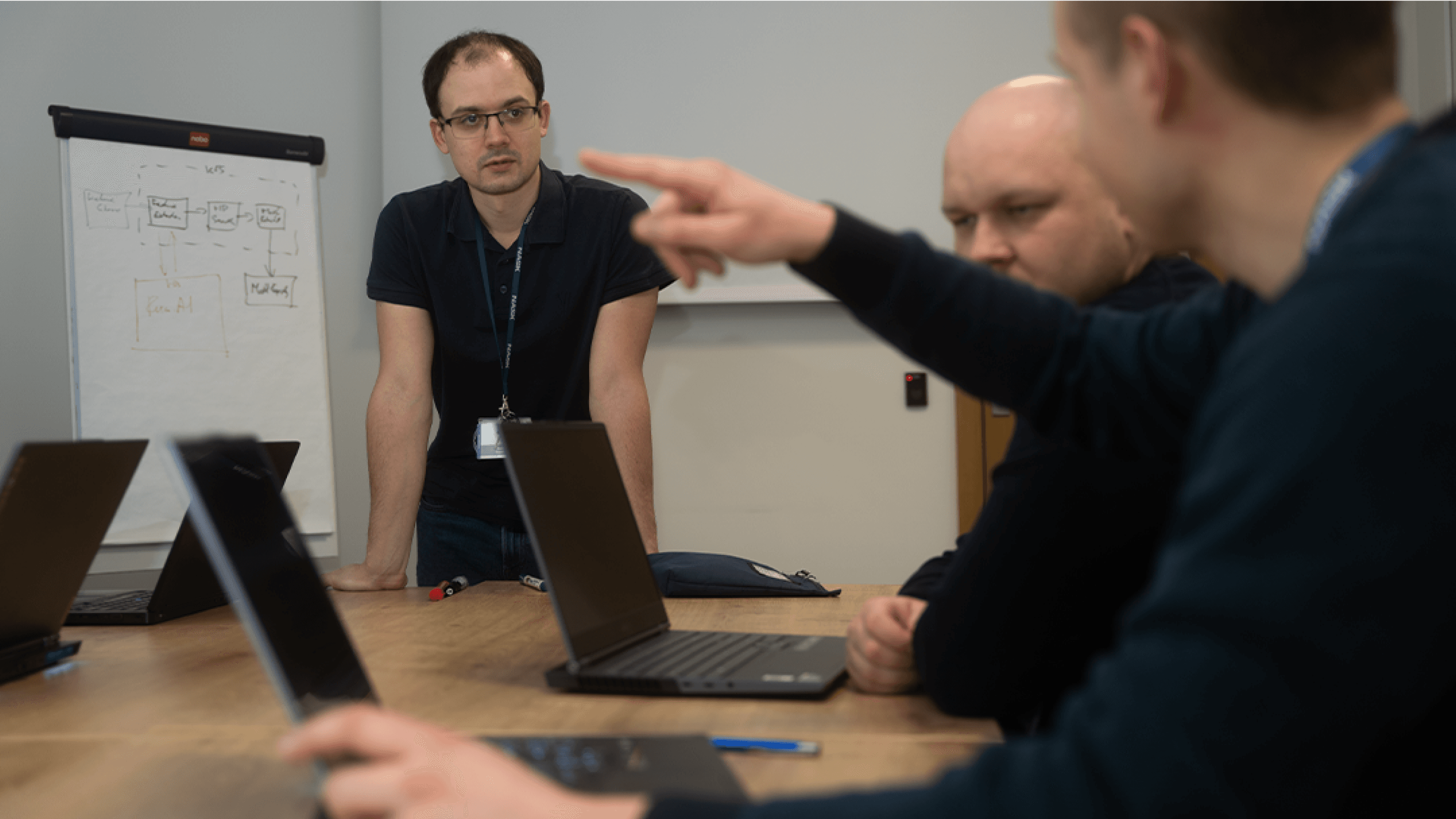 Trzech naukowców - dwóch siedzi przy stole, jeden stoi. Na stole znajdują się komputery.