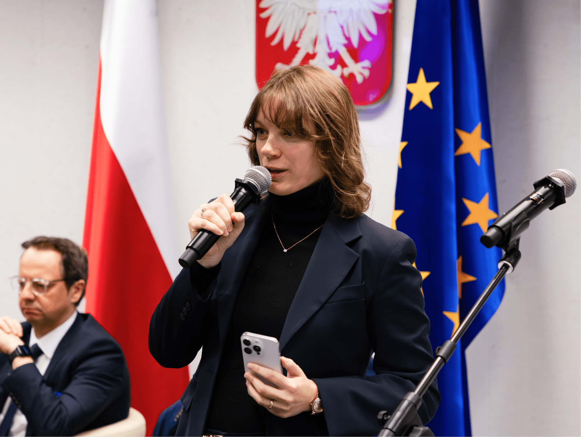 Kobieta zabierająca głos podczas konferencji. W tle godło Polski, flaga Polki i Unii Europejskiej.