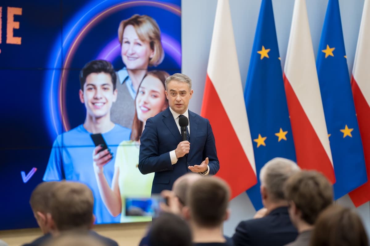 Mężczyzna w garniturze przemawia do publiczności, trzymając mikrofon. W tle widać flagi Polski i Unii Europejskiej oraz ekran z grafiką przedstawiającą uśmiechniętych młodych ludzi i kobietę w średnim wieku.