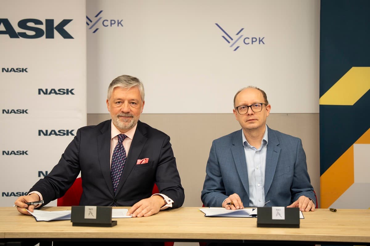 Dwaj mężczyźni siedzą przy stole i podpisują dokument o współpracy między NASK a CPK. Po lewej stronie siedzi prezes CPK w granatowym garniturze, uśmiechnięty, z długopisem w dłoni. Po prawej stronie znajduje się dyrektor NASK, dr inż. Radosław Nielek, w jasnoniebieskiej marynarce. Obaj podpisują dokument o współpracy przy stole, a w tle widoczne są logotypy NASK i CPK.